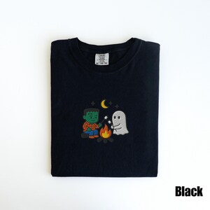 May include: Black t-shirt with an embroidered design featuring a Frankenstein monster and a ghost roasting marshmallows over a campfire. The design includes a crescent moon and stars. The text "Black" is in the bottom right corner.