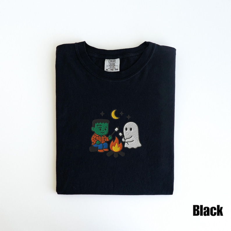 May include: Black t-shirt with an embroidered design featuring a Frankenstein monster and a ghost roasting marshmallows over a campfire. The design includes a crescent moon and stars. The text "Black" is in the bottom right corner.