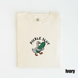 May include: Ivory-colored t-shirt with the phrase "PICKLE SLUT" above an embroidered cartoon duck holding a pickle. The shirt is folded and has a tag that says "COMFORT COLORS".