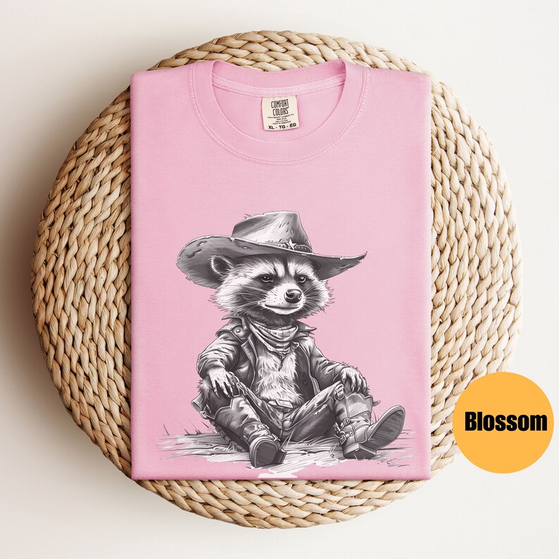 May include: Pink t-shirt with a black and white illustration of a raccoon wearing a cowboy hat and a leather jacket. The raccoon is sitting with its legs crossed and wearing boots. The text "COMFORT COLORS" is visible on the tag.