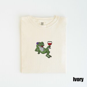 Pu&ograve; includere: T-shirt color avorio con un disegno ricamato di una rana verde con un fiore viola e un bicchiere di vino rosso. La rana ha il rossetto rosso. La maglietta &egrave; piegata.