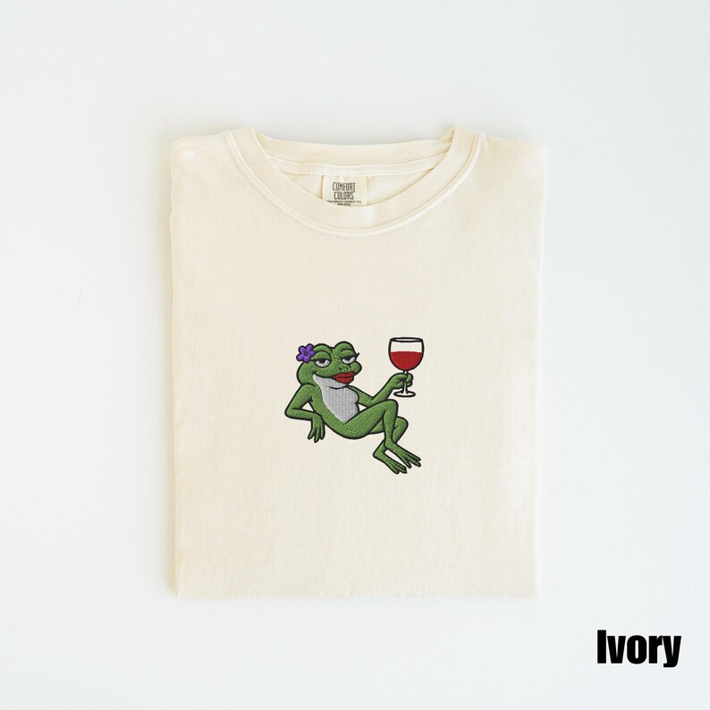 Pu&ograve; includere: T-shirt color avorio con un disegno ricamato di una rana verde con un fiore viola e un bicchiere di vino rosso. La rana ha il rossetto rosso. La maglietta &egrave; piegata.