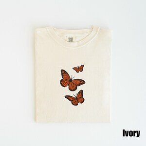 May include: Ivory-colored t-shirt with three embroidered orange butterflies. The butterflies are in different sizes and arranged vertically. The shirt is folded neatly and has a tag at the neckline. The word "Ivory" is printed at the bottom right.