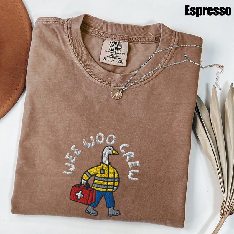May include: Espresso-colored t-shirt with the embroidered phrase "WEE WOO CREW" and a cartoon duck carrying a red first aid kit. The shirt has a crew neck and a Comfort Colors tag. A silver necklace with a small pendant is draped across the shirt.