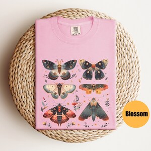 Puede incluir: Camiseta rosa con un gr&aacute;fico de seis polillas coloridas con detalles intrincados. Las polillas est&aacute;n dispuestas en una cuadr&iacute;cula con flores y hojas que las rodean. El texto "COMFORT COLORS" est&aacute; impreso en la camiseta.