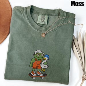 May include: Moss green t-shirt with an embroidered design of a frog and a duck on a skateboard. The frog wears glasses, a hat, and orange overalls. The duck wears a blue hat. A gold necklace with a pendant is on the shirt.