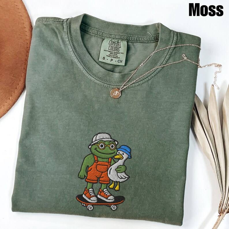 May include: Moss green t-shirt with an embroidered design of a frog and a duck on a skateboard. The frog wears glasses, a hat, and orange overalls. The duck wears a blue hat. A gold necklace with a pendant is on the shirt.