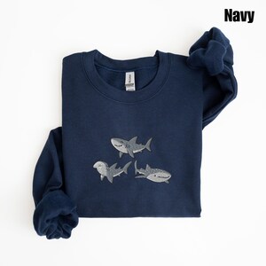 May include: Navy blue sweatshirt featuring embroidered shark designs. The sweatshirt has a crew neck and long sleeves. The embroidered sharks are in shades of gray and white. The word "Navy" is printed in the upper right corner.