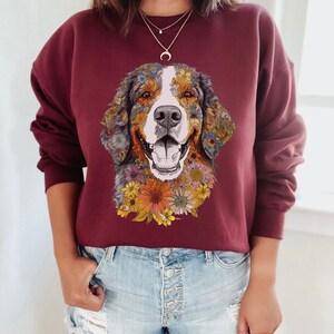 K&ouml;nnte beinhalten: Ein maroonfarbener Pullover mit einem Aufdruck eines Berner Sennenhundes, dessen Gesicht von bunten Blumen umgeben ist. Der Hund l&auml;chelt und hat eine wei&szlig;e Schnauze.