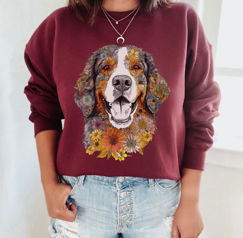 K&ouml;nnte beinhalten: Ein maroonfarbener Pullover mit einem Aufdruck eines Berner Sennenhundes, dessen Gesicht von bunten Blumen umgeben ist. Der Hund l&auml;chelt und hat eine wei&szlig;e Schnauze.
