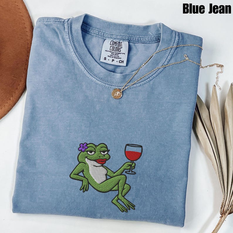 Pu&ograve; includere: Una maglietta blu jeans Comfort Colors con un ricamo di una rana verde che tiene in mano un calice di vino. La rana ha un fiore viola e rossetto rosso. Una collana d'oro con un ciondolo a forma di rosa &egrave; sulla maglietta.