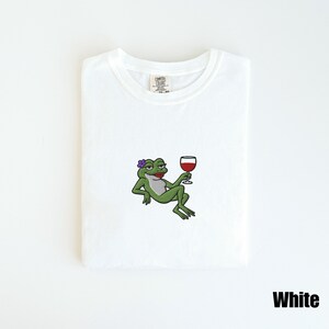 Pu&ograve; includere: T-shirt bianca con un ricamo di una rana verde con un fiore viola, rossetto rosso e un bicchiere di vino rosso. La maglietta ha uno scollo tondo e la scritta "White" &egrave; in basso a destra.