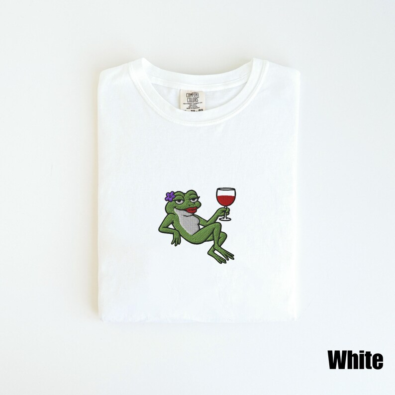 Pu&ograve; includere: T-shirt bianca con un ricamo di una rana verde con un fiore viola, rossetto rosso e un bicchiere di vino rosso. La maglietta ha uno scollo tondo e la scritta "White" &egrave; in basso a destra.