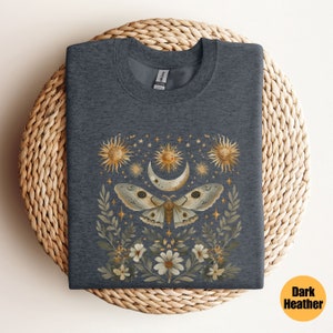 Pode incluir: Camiseta cinza escuro mesclada com um gr&aacute;fico de uma mariposa com asas douradas, cercada por uma lua crescente, estrelas e flores.