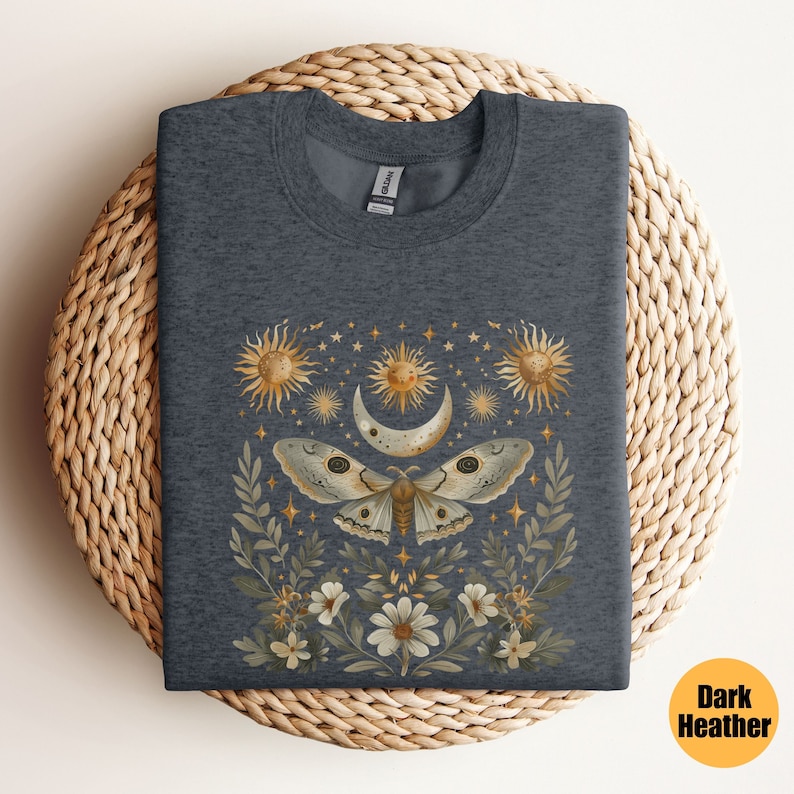 Pode incluir: Camiseta cinza escuro mesclada com um gr&aacute;fico de uma mariposa com asas douradas, cercada por uma lua crescente, estrelas e flores.