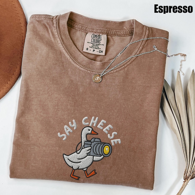 May include: Espresso-colored t-shirt with a crew neck and short sleeves. The shirt features an embroidered design of a goose holding a camera, with the words "Say Cheese" above the goose. A silver necklace with a small pendant is draped across the shirt.