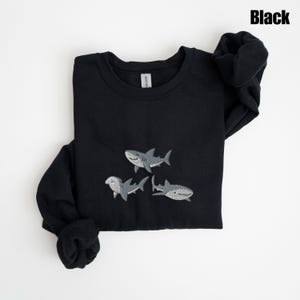 May include: Black sweatshirt featuring three embroidered gray sharks. The word "Black" is printed in the upper right corner. The sweatshirt is folded, with the sleeves and cuffs visible.