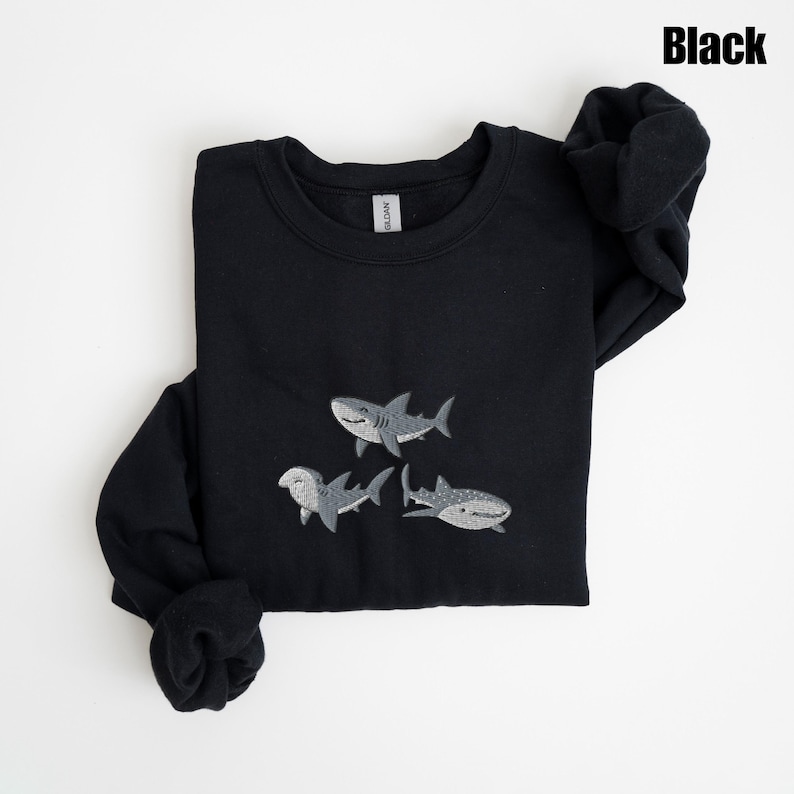 May include: Black sweatshirt featuring three embroidered gray sharks. The word "Black" is printed in the upper right corner. The sweatshirt is folded, with the sleeves and cuffs visible.