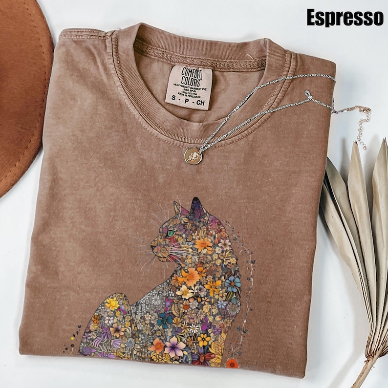 May include: A brown t-shirt with a floral cat design. The shirt is made of a soft, comfortable material and has a distressed look. The text "COMFORT COLORS" is printed on the tag.