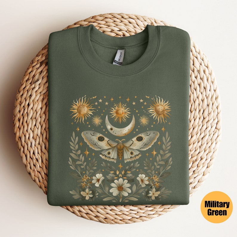 Pode incluir: Uma camisa de moletom verde com uma estampa de uma mariposa, lua e estrelas em dourado e branco. A estampa &eacute; cercada por flores e folhas.
