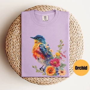 May include: A lavender t-shirt featuring a colourful bird and floral design. The bird is blue, orange, and yellow, and the flowers are pink, yellow, and blue. The shirt has the text "COMFORT COLORS" on the tag.