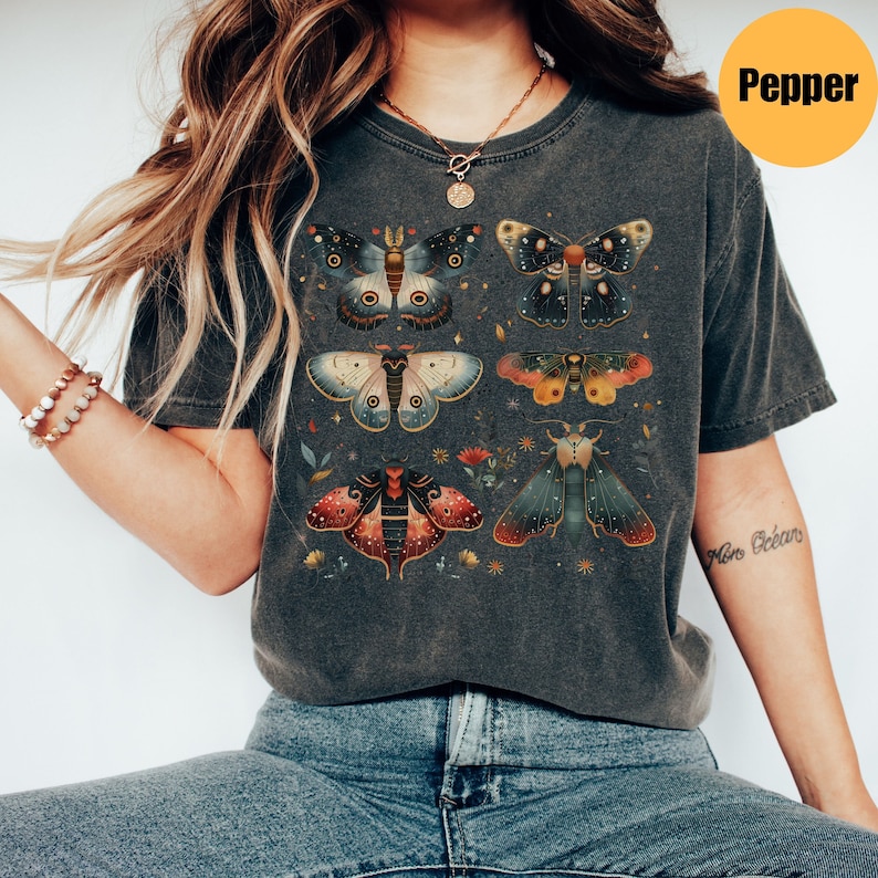 Puede incluir: Una camiseta gris oscuro con un estilo vintage que presenta seis ilustraciones coloridas de mariposas y polillas. La camiseta tiene cuello redondo y mangas cortas.