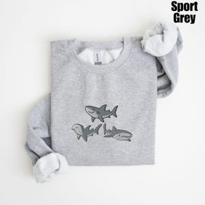 May include: Sport Grey sweatshirt featuring three cartoon sharks in shades of gray. The sharks are smiling and have detailed fins and markings. The sweatshirt has a crew neck and long sleeves with a soft, white interior.