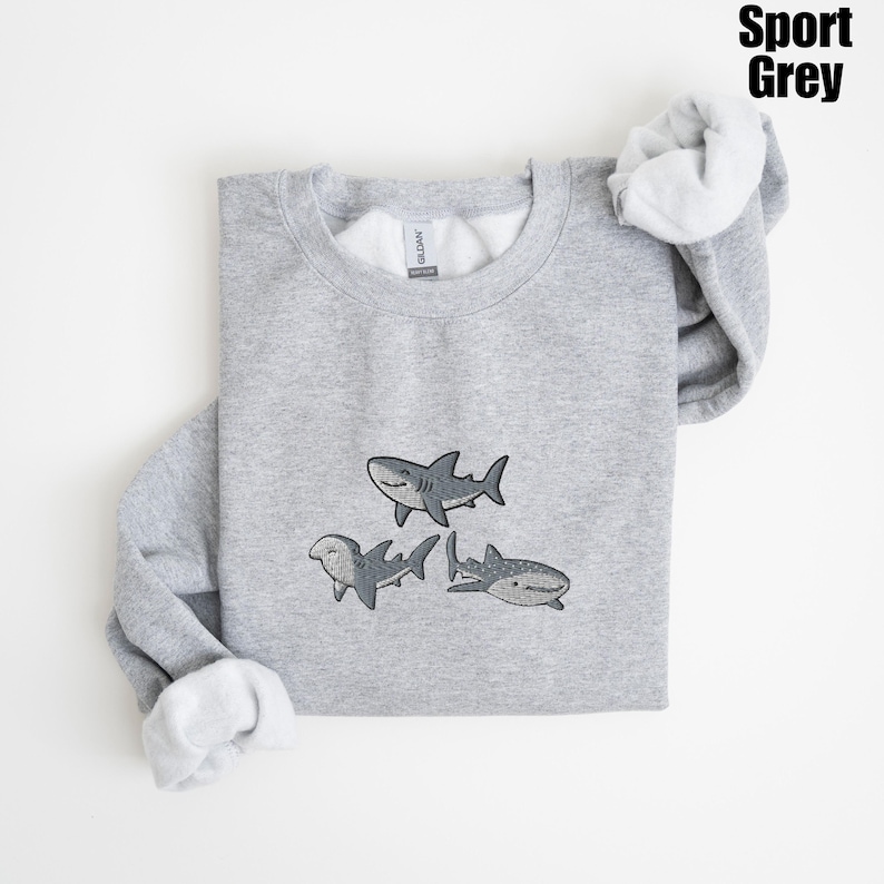 May include: Sport Grey sweatshirt featuring three cartoon sharks in shades of gray. The sharks are smiling and have detailed fins and markings. The sweatshirt has a crew neck and long sleeves with a soft, white interior.