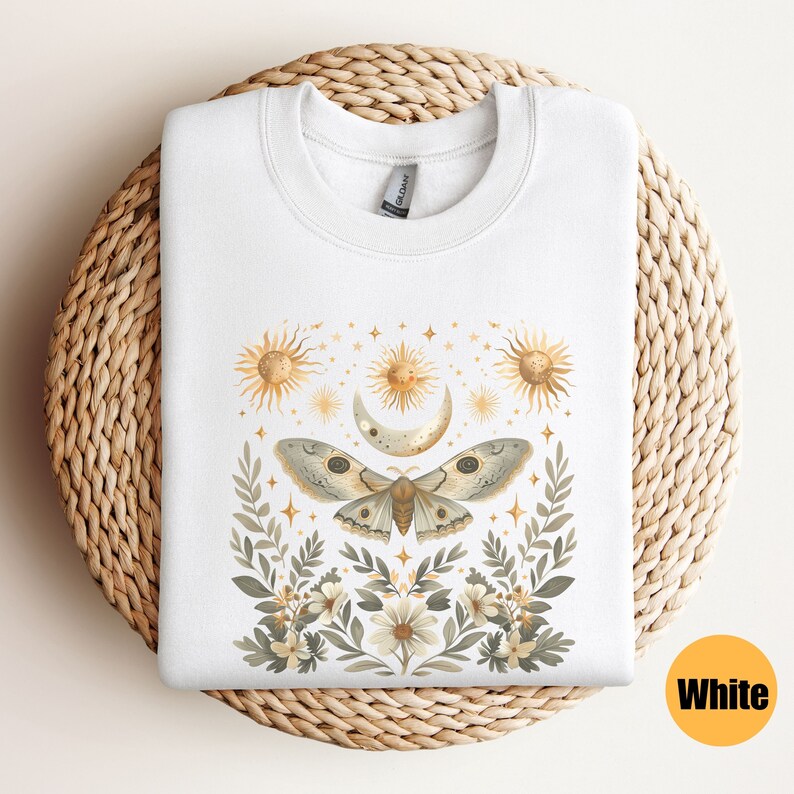 Pode incluir: Camisola branca com um design gr&aacute;fico de uma mariposa com as asas abertas, rodeada de flores, luas e estrelas. O design &eacute; em tons de dourado, castanho e branco.