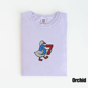 Peut inclure: T-shirt Comfort Colors violet clair avec un motif brod&eacute; d'une oie tenant les chiffres 6 et 7. Le t-shirt est &eacute;tiquet&eacute; "Orchid" en bas &agrave; droite.