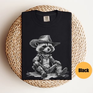 May include: Black t-shirt with a graphic of a raccoon wearing a cowboy hat and leather jacket. The raccoon is sitting with its legs crossed and wearing cowboy boots.