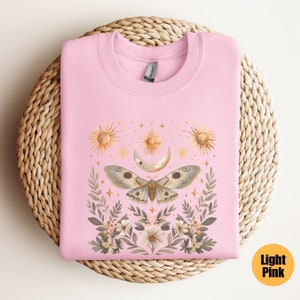 Pode incluir: Uma camisa de cor rosa claro com um design gr&aacute;fico de uma mariposa com as asas abertas, cercada por flores, luas e estrelas. O design &eacute; em tons de dourado, marrom e branco.