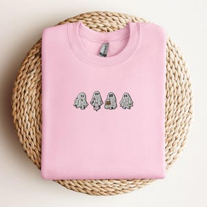 May include: A light pink crewneck sweatshirt featuring an embroidered design of four small white ghosts in a horizontal row. Each ghost has a unique pattern, including floral and pumpkin motifs, stitched in various colours on the chest area.