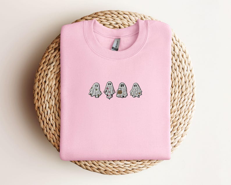 May include: A light pink crewneck sweatshirt featuring an embroidered design of four small white ghosts in a horizontal row. Each ghost has a unique pattern, including floral and pumpkin motifs, stitched in various colours on the chest area.