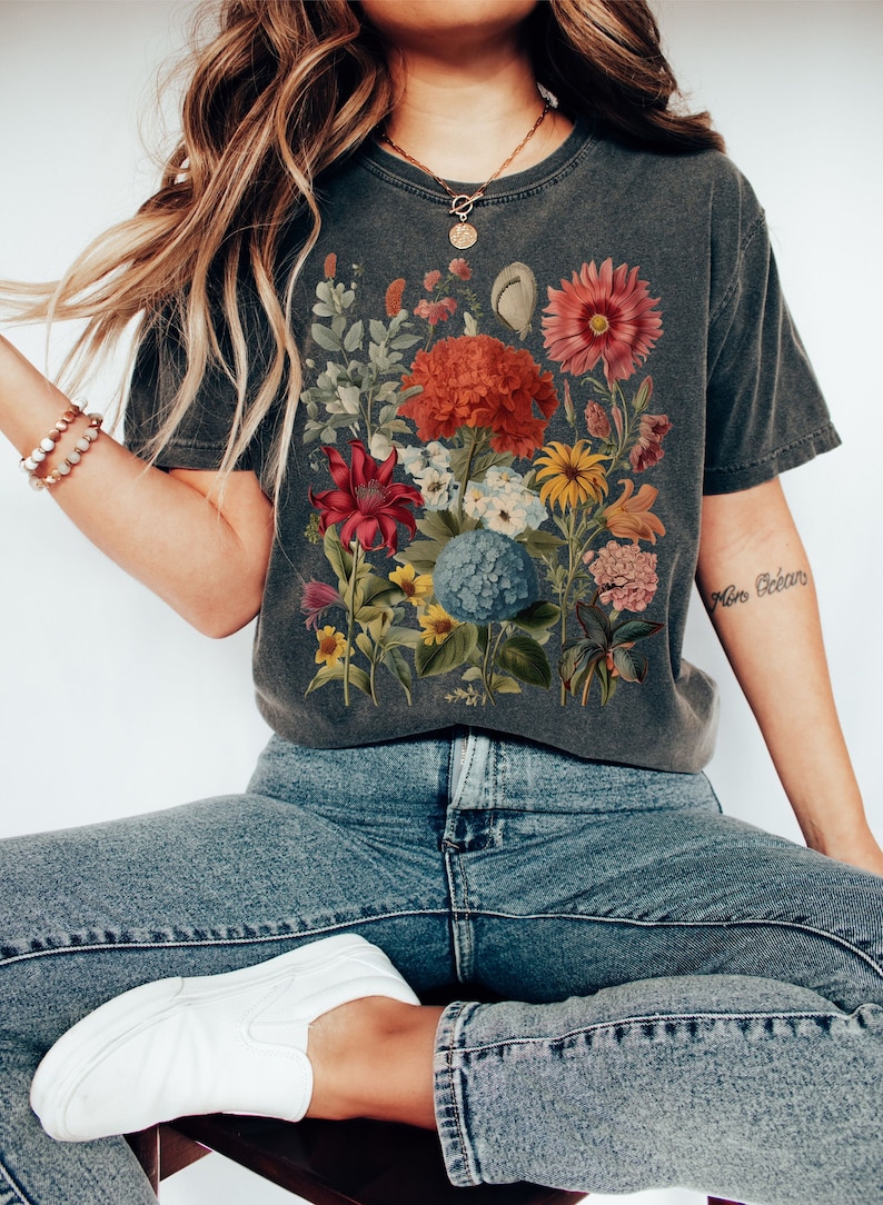 Pu&ograve; includere: Una maglietta grigia scura con una stampa floreale che presenta una variet&agrave; di fiori in tonalit&agrave; di rosso, rosa, giallo, blu e verde. I fiori sono disposti in un design simile a un bouquet.