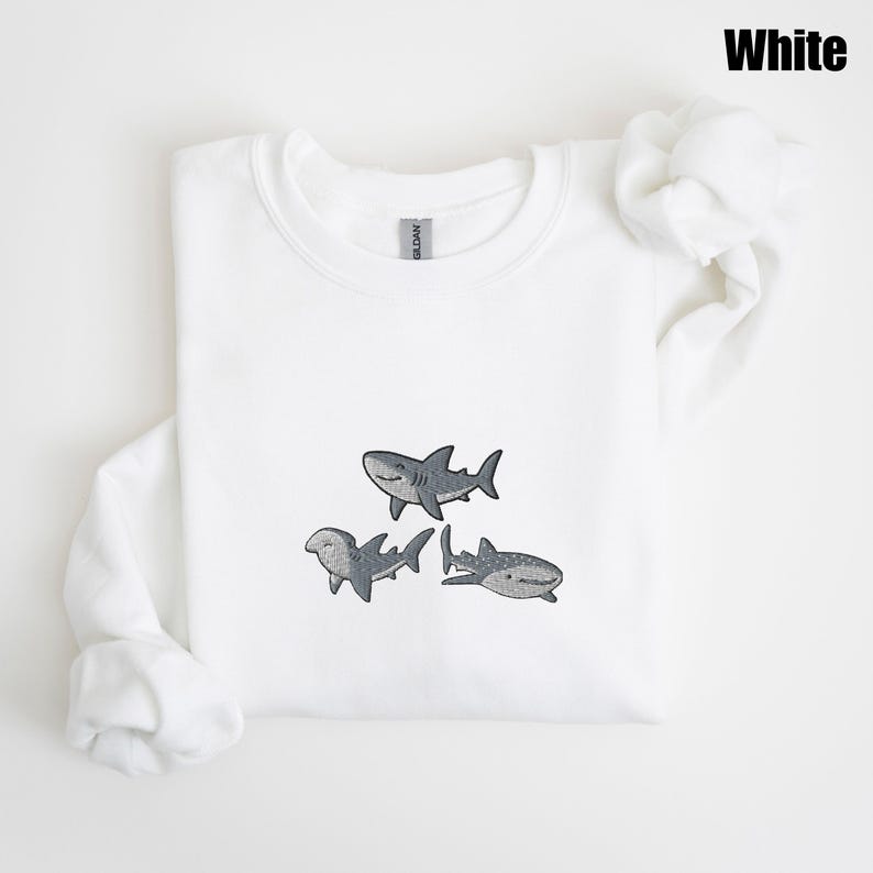 May include: White sweatshirt featuring three embroidered gray sharks. The sharks have detailed fins and smiling faces. The word "White" is printed in black in the upper right corner. The sweatshirt is made of a soft material.