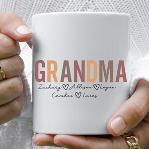 以下が含まれることがあります： オレンジ、ブラウン、ピンクの文字で「GRANDMA」と書かれた白いセラミックマグカップ。単語の下には、各名前の間にハートマークが付いた「Zachary Allison Logan Camden Lucas」と書いてあります。