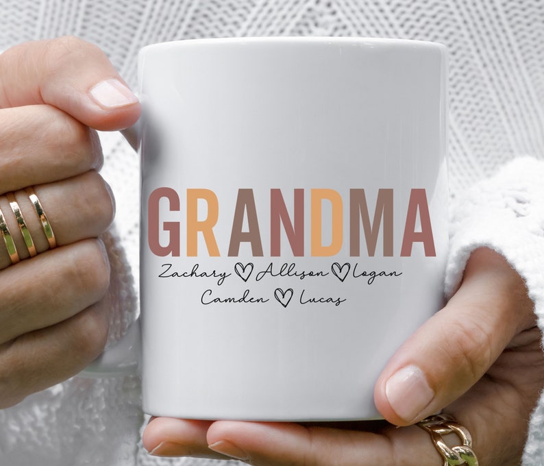 以下が含まれることがあります： オレンジ、ブラウン、ピンクの文字で「GRANDMA」と書かれた白いセラミックマグカップ。単語の下には、各名前の間にハートマークが付いた「Zachary Allison Logan Camden Lucas」と書いてあります。