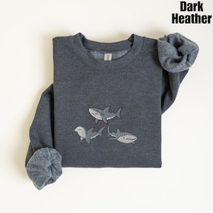 May include: Dark heather gray sweatshirt with three embroidered shark designs. The sharks are in shades of gray and white. The sweatshirt has a crew neck and long sleeves. The text "Dark Heather" is in the upper right corner.