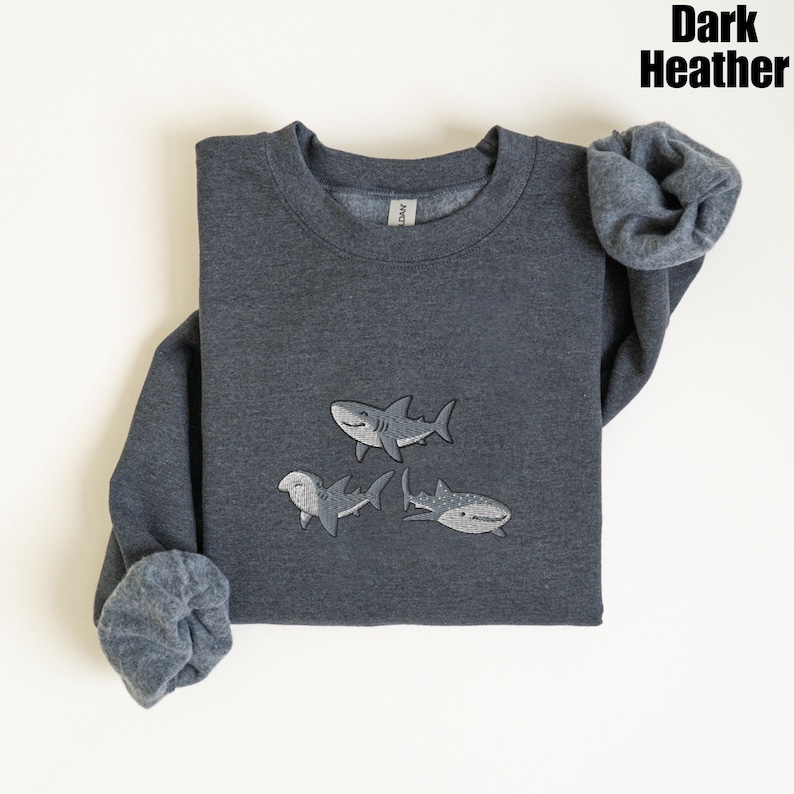 May include: Dark heather gray sweatshirt with three embroidered shark designs. The sharks are in shades of gray and white. The sweatshirt has a crew neck and long sleeves. The text "Dark Heather" is in the upper right corner.