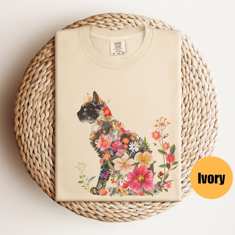 May include: A beige t-shirt featuring a floral cat design. The cat is composed of colourful flowers and is sitting on a white background. The shirt is made from 100% cotton and is available in sizes S-XL. The text "COMFORT COLORS" is printed on the tag.