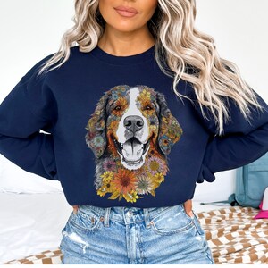 K&ouml;nnte beinhalten: Ein marineblaues Sweatshirt mit einem farbenfrohen Blumendesign eines Berner Sennenhunds. Das Gesicht des Hundes ist von Blumen in Gelb-, Orange-, Rosa- und Lilat&ouml;nen umgeben.