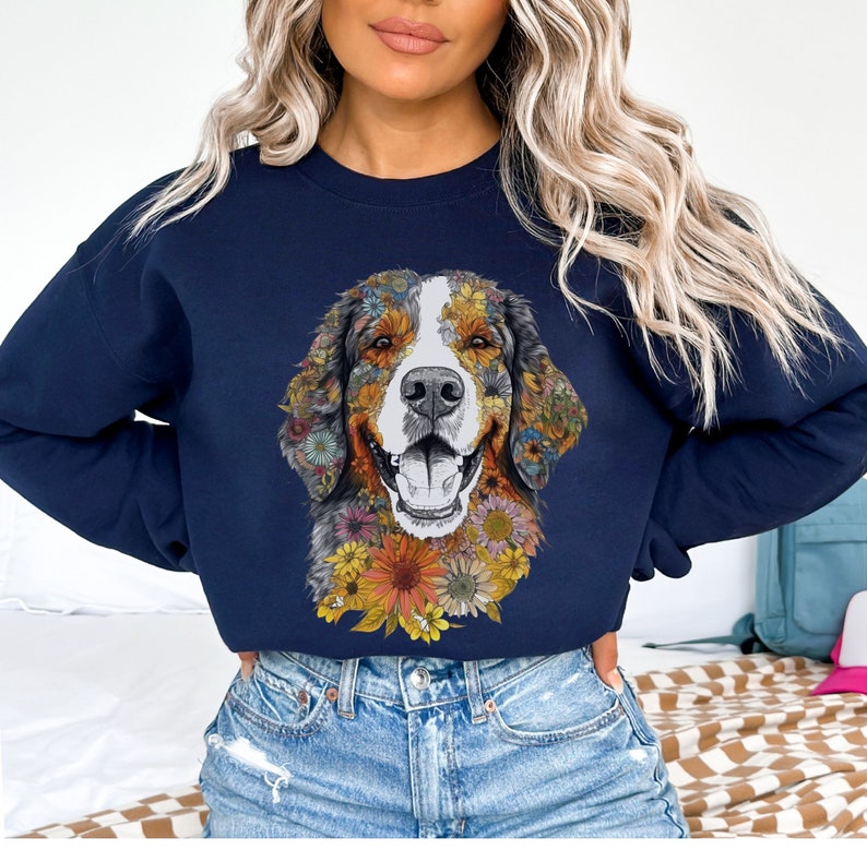 K&ouml;nnte beinhalten: Ein marineblaues Sweatshirt mit einem farbenfrohen Blumendesign eines Berner Sennenhunds. Das Gesicht des Hundes ist von Blumen in Gelb-, Orange-, Rosa- und Lilat&ouml;nen umgeben.