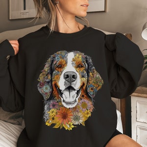 K&ouml;nnte beinhalten: Schwarzes Sweatshirt mit einem farbenfrohen Blumendesign eines Berner Sennenhunds. Das Gesicht des Hundes ist von Blumen in Gelb-, Orange-, Rot- und Rosat&ouml;nen umgeben.