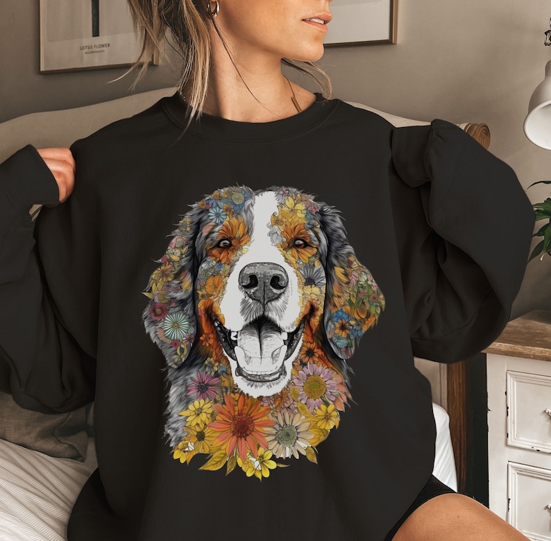 K&ouml;nnte beinhalten: Schwarzes Sweatshirt mit einem farbenfrohen Blumendesign eines Berner Sennenhunds. Das Gesicht des Hundes ist von Blumen in Gelb-, Orange-, Rot- und Rosat&ouml;nen umgeben.