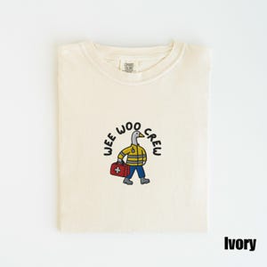 May include: Ivory-colored t-shirt with a cartoon duck wearing a yellow jacket and blue pants, carrying a red medical bag. The text "WEE WOO CREW" is arched above the duck. The shirt is folded neatly.