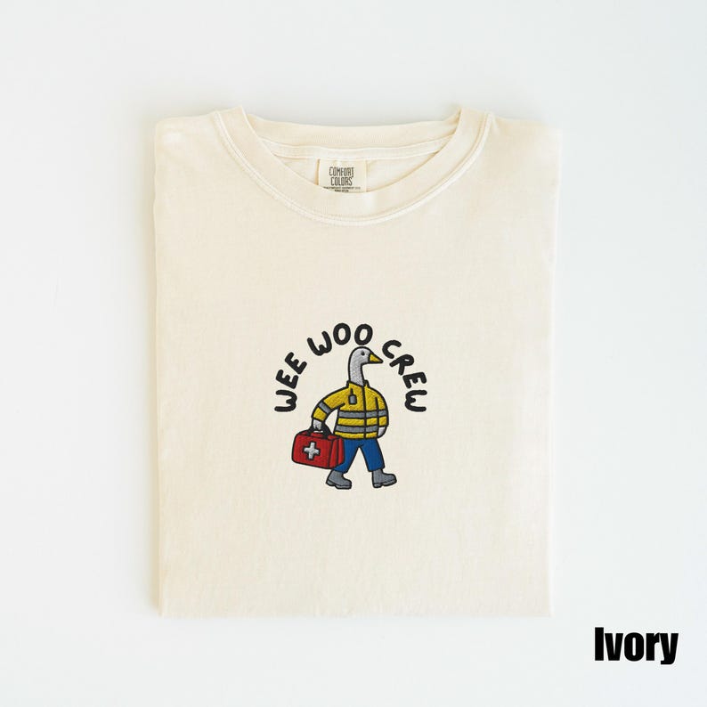 May include: Ivory-colored t-shirt with a cartoon duck wearing a yellow jacket and blue pants, carrying a red medical bag. The text "WEE WOO CREW" is arched above the duck. The shirt is folded neatly.