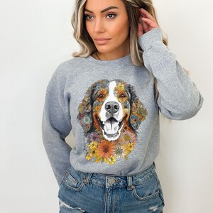 K&ouml;nnte beinhalten: Ein graues Sweatshirt mit einem floralen Design eines Berner Sennenhunds. Das Fell des Hundes besteht aus bunten Blumen und der Hintergrund ist einfarbig grau.