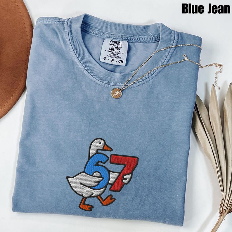Peut inclure: T-shirt bleu jean Comfort Colors avec un motif brod&eacute; d'une oie portant le num&eacute;ro 67. Un collier en or avec un pendentif est pos&eacute; sur le t-shirt. Le t-shirt est de taille S-P-CH.