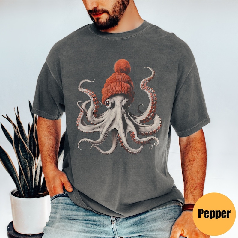 May include: Dark gray t-shirt featuring a detailed illustration of an octopus wearing a red knit beanie with a pom-pom. The octopus has white skin, red suckers, and large eyes. The shirt is a casual style.